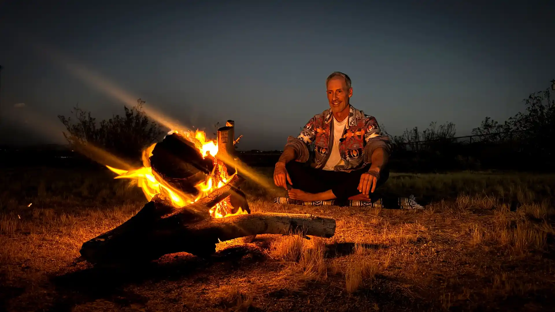 Man relaxing beside a glowing campfire under the evening sky at a Joshua Tree desert retreat.